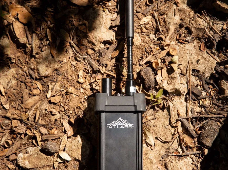 AT Labs RM-2 ruggedized mesh radio on rocky ground, antenna up, AT Labs logo visible on the housing